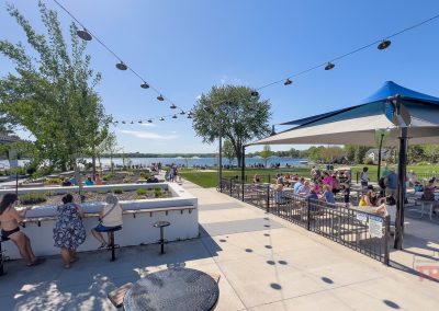 People gather at Antlers Park in Lakeville MN, enjoying picnic tables, string lights, and lakeside views on a sunny day. Some relax under umbrellas while others lounge on the grass near the water’s edge.