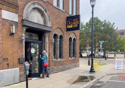A person stands outside the entrance of B-Side Cafe on a Saint Cloud MN street corner; the brick building has arched windows and the wet sidewalk is lined with trees and street signs in the background.