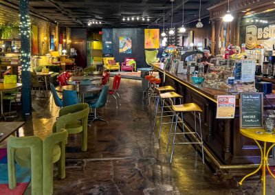 A cozy, colorful spot in Saint Cloud MN, B-Side Cafe features eclectic furniture, vibrant decor, artwork on brick walls, string lights, and a long wooden counter with pastries and drinks. A staff member stands behind the counter.