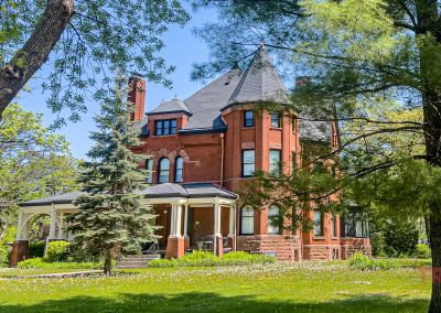 A Victorian red brick mansion with a prominent turret and wraparound porch sits amidst lush green lawns and mature trees under a clear blue sky in St. Cloud MN.