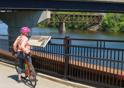 A person wearing a pink helmet and orange jacket stands with a bicycle on the Beaver Island Trail in St. Cloud MN, reading an informational sign beside a river, with bridges and green trees in the background.