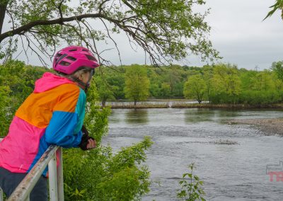 A person in a pink helmet and colorful jacket leans on a railing, looking out over the river along the scenic Beaver Island Trail in St. Cloud MN, surrounded by green trees on a cloudy day.