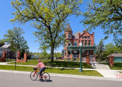 A person rides a bicycle past a large, ornate red-brick Victorian house with a turret and wraparound porch on a sunny day in St. Cloud MN, surrounded by lush green trees and grass.