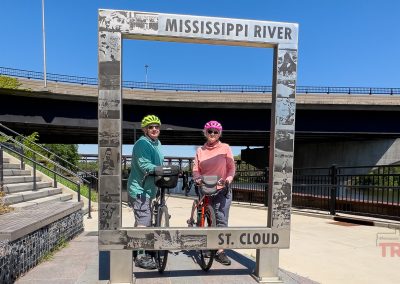 Two cyclists wearing helmets pose with their bikes inside a metal frame labeled Mississippi River and St. Cloud MN, near the scenic Beaver Island Trail, with a bridge and stairs visible in the background on a sunny day.