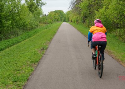 A person wearing a pink and orange jacket and helmet rides a bicycle down the paved Beaver Island Trail in St. Cloud MN, surrounded by green foliage on an overcast day. The tree-lined path stretches straight ahead.