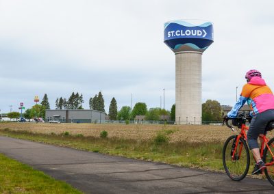 A person in a pink jacket and helmet rides a bicycle on the Beaver Island Trail toward a large water tower labeled ST. CLOUD, with buildings and signs visible in the background near St. Cloud MN.