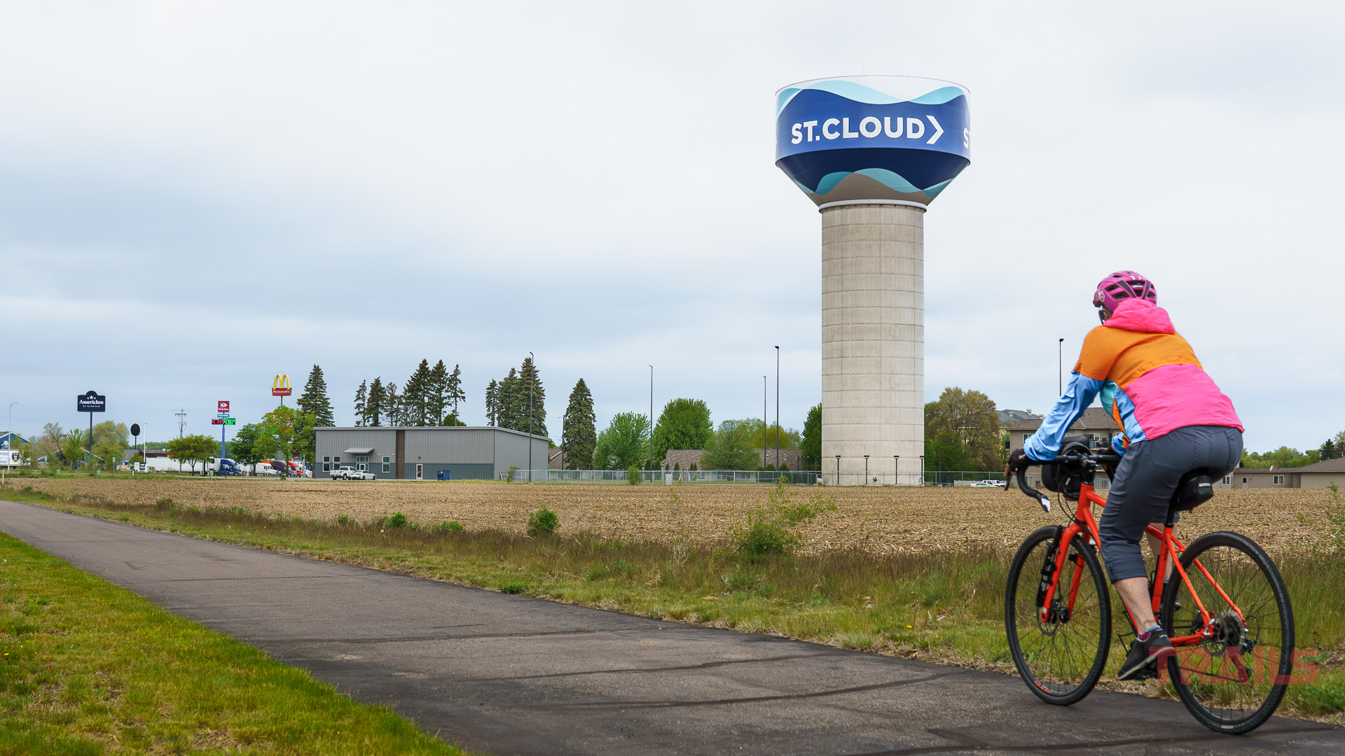 Beaver Island Trail Saint Cloud A cyclist in a pink jacket rides along the Beaver Island Trail toward a large water tower labeled ST. CLOUD, with businesses and trees in the background under a cloudy St. Cloud MN sky.