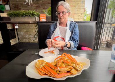 A person wearing glasses and a bib sits at a Boil Seafood Restaurant booth in Saint Cloud MN, cracking crab legs by hand. A large plate of crab legs is on the table, with natural light streaming through windows in the background.