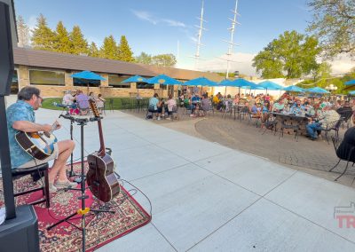 A musician plays guitar and sings on an outdoor patio at Brianno's Chart House in Lakeville, MN, as people dine under blue umbrellas. Trees and a stone building are in the background on a sunny day.