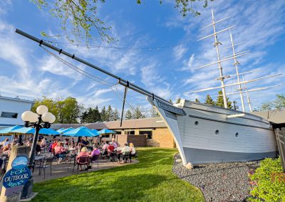 People dine outdoors on a sunny day beside blue umbrellas and a large ship replica built against Brianno's Chart House in Lakeville, MN. A chalkboard sign and lamp post are in the foreground; the sky is bright with scattered clouds.