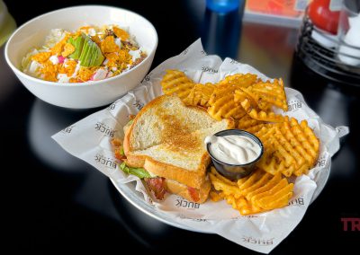 A plate with a toasted sandwich, waffle fries, and ranch dip sits next to a fresh salad with avocado, corn, cheese, and tomatoes on a dark table—perfect for a Buck Hill lunch.