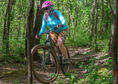 A person wearing a pink helmet, sunglasses, blue long-sleeve shirt, and colorful shorts rides a mountain bike along the Buck Hill mountain bike trails, winding through a lush, green forest as sunlight filters through the trees.