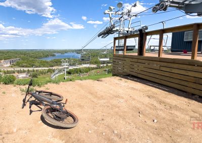 A mountain bike rests on a dirt trail near a wooden deck and ski lift, overlooking the scenic Buck Hill mountain bike trails, with trees, a lake, and buildings under a sunny blue sky.
