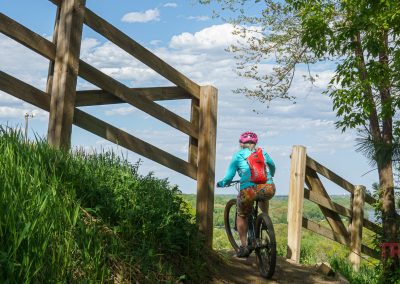 A cyclist wearing a pink helmet and colorful clothes rides a mountain bike through a wooden gate on the lush green Buck Hill mountain bike trails under a partly cloudy sky.