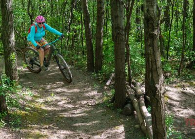 A person wearing a pink helmet and blue shirt rides a mountain bike along the narrow Buck Hill mountain bike trails through a green, wooded forest. Sunlight filters through the trees.
