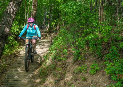 A person wearing a pink helmet and blue jacket rides a mountain bike along the narrow Buck Hill mountain bike trails, weaving through a lush, green forest as sunlight filters through the trees and dances on the wooden path.