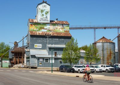 A person on a bicycle rides past the Farmington grain elevator, featuring a mural reading LIFE: THE GIFT OF EARTH. Several silos, parked cars, and green trees are visible in this small town under a clear blue sky.