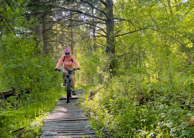 A person wearing a pink helmet and shirt rides a mountain bike along the Jail Trail, a narrow wooden path winding through Saint Cloud MN’s lush, green forest on a sunny day.