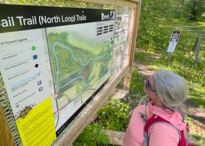 A woman with gray hair and a pink backpack reads a large trail map on a sign labeled Jail Trail (North Loop) Trails at the edge of a wooded path in Saint Cloud, MN. Sunlight filters through the trees in the background.