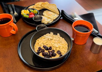 A breakfast table at Jules' Bistro Saint Cloud features a bowl of oatmeal with sliced almonds and dried cranberries, two orange mugs of coffee, toast, eggs, mixed fruit, and utensils on black napkins.