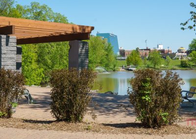 A lakeside park in Saint Cloud MN with a wooden pergola, stone pillars, trimmed bushes, benches, a fountain in Lake George, and buildings visible in the background under a clear blue sky.