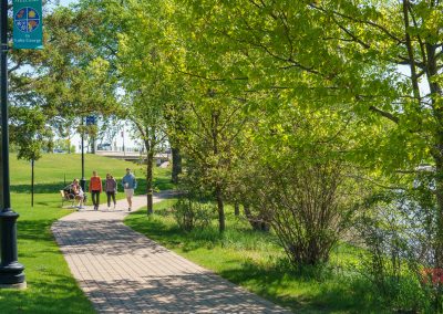 A paved park path curves through green trees and grass in Saint Cloud MN. Four people walk together while two others sit on a bench. A lamppost displays a Lake County sign, and sunlight filters through the leaves near Lake George.