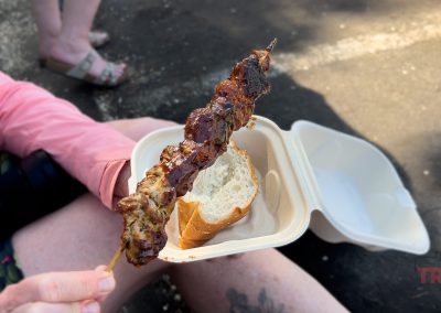 A person seated outdoors holds a takeout container with a piece of bread and a grilled meat skewer, likely from Lakeville Food Truck Fest. Another person’s legs are visible in the background on a paved surface.