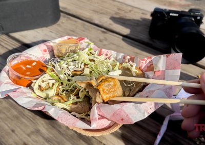 A hand holding chopsticks lifts a dumpling from a basket filled with dumplings, shredded cabbage, and orange dipping sauce at the Lakeville Food Truck Fest, on a wooden table with a camera and case in the background.