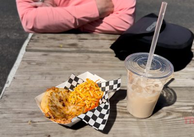 A wooden table at Lakeville Food Truck Fest holds a tray with a stuffed tortilla and slaw, beside a plastic cup of iced drink. An arm in a pink sleeve rests on the table, with a black bag nearby.