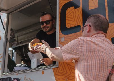 A man wearing sunglasses hands food in a paper tray to another man at a food truck window during the Lakeville Food Truck Fest. The customer reaches out for the meal as bold orange graphics decorate the vibrant food truck exterior.