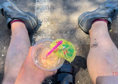 A person sits on asphalt with legs outstretched, holding an iced drink with a pink straw and decorative umbrella at the Lakeville Food Truck Fest. They wear biking shoes and red socks; a camera lens lies between their legs.