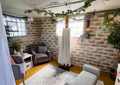 A cozy room with exposed brick walls, armchairs, a rug, potted plants, and string lights in Lavender Barnyard, Farmington MN. A long white dress hangs in the center as sunlight streams through sheer curtains amid leafy decor.