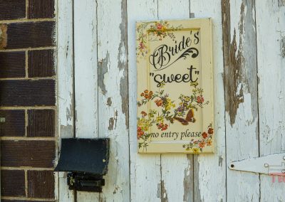 A decorative sign reading Brides Sweet—no entry please hangs on a weathered white wooden door at Lavender Barnyard in Farmington MN, with flowers on the sign. The door stands by a brown brick wall and a black mailbox.
