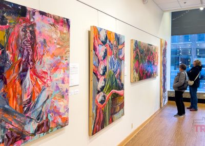 Two people view colorful, abstract paintings in a bright art gallery at the Paramount Theater, with large windows overlooking the bustling streets of Saint Cloud MN.