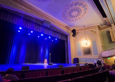 A theater stage at the Paramount Theater in Saint Cloud MN, with a stool and microphone under blue lights, ornate ceiling, and audience members seated and waiting for a performance to begin.