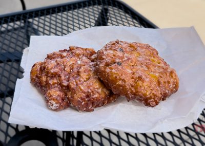 Two glazed apple fritters from Sweet Kneads Bakery rest on white parchment paper, placed on a black metal outdoor table in Farmington MN.