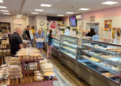 Customers stand in line and browse pastries at Sweet Kneads Bakery, a bright spot in Farmington MN, with glass display cases full of baked goods. Shelves hold extra treats while staff assist customers, all surrounded by colorful wall art.