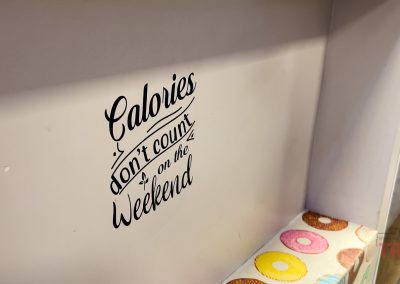 A wall at Sweet Kneads Bakery in Farmington MN displays the quote "Calories don't count on the Weekend" in decorative black text, with a colorful donut-patterned surface visible below.