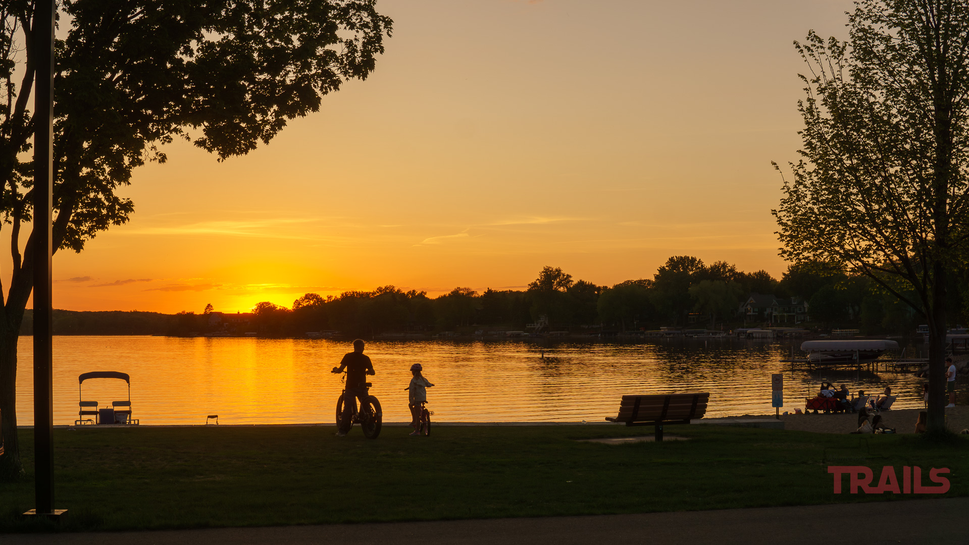 The Lakeville Life Two people ride bikes along the Antlers Park lakeshore at sunset, with trees framing the scene, benches nearby, and boats on the calm water. The sky glows orange and yellow over Lakeville MN, creating a peaceful atmosphere.