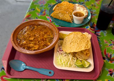 A tray with a bowl of soup, two crispy tacos on cabbage with lime wedges and onions, and a blue spoon. In the background, another plate with rice, beans, tacos, salsa, and a drink sits on a colorful table in Saint Cloud MN.