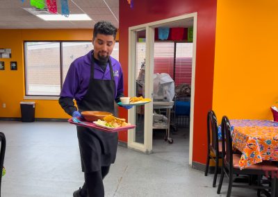 A server wearing a purple shirt and black apron carries two trays of food in a brightly colored Saint Cloud MN restaurant with orange and red walls and patterned tablecloths.