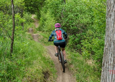 A person wearing a pink helmet and red backpack rides a bicycle on a narrow dirt trail at West Lake Marion Mountain Bike Trails in Lakeville, MN, surrounded by lush green trees and vegetation.