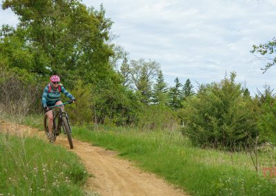 A person wearing a pink helmet and striped shirt rides a mountain bike down a dirt trail at West Lake Marion Mountain Bike Trails in Lakeville, MN, surrounded by green grass, trees, and bushes under a partly cloudy sky.