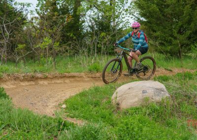 A person in a pink helmet and blue patterned jacket rides a mountain bike around a dirt trail curve at West Lake Marion Mountain Bike Trails in Lakeville, MN, surrounded by green woods, grass, bushes, and rocks.