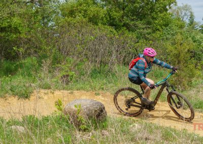 A cyclist wearing a pink helmet and blue striped shirt rides a mountain bike on the West Lake Marion Mountain Bike Trails in Lakeville MN, curving around a large rock amid green bushes and trees.