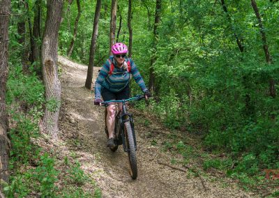 A person wearing a pink helmet and colorful jacket rides a mountain bike along a narrow dirt trail through the dense, green forest of West Lake Marion Mountain Bike Trails in Lakeville, MN.