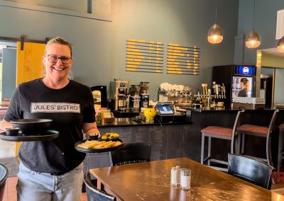 A smiling server at Jules’ Bistro in Saint Cloud MN holds plates of food inside a cozy restaurant with modern decor, pendant lighting, and a coffee counter in the background.