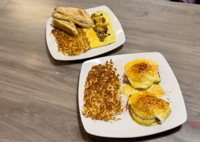 Two plates of breakfast food on a wooden table at Bader’s Café in Albert Lea MN: one with eggs Benedict, hash browns, and sauce; the other with toast, scrambled eggs, hash browns, and sausage gravy.