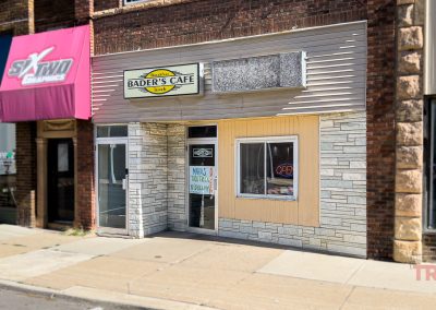 A small cafe with a sign reading Bader’s Café is nestled on a city sidewalk in Albert Lea MN. An open sign hangs in the window and signs are displayed on the door. Storefronts are visible on either side.