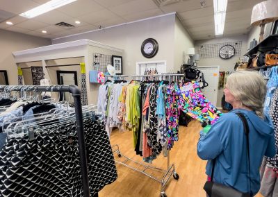 A woman with gray hair browses colorful clothing on racks inside Between Friends Boutique in Albert Lea, MN, where wooden floors and various garments are beautifully displayed.